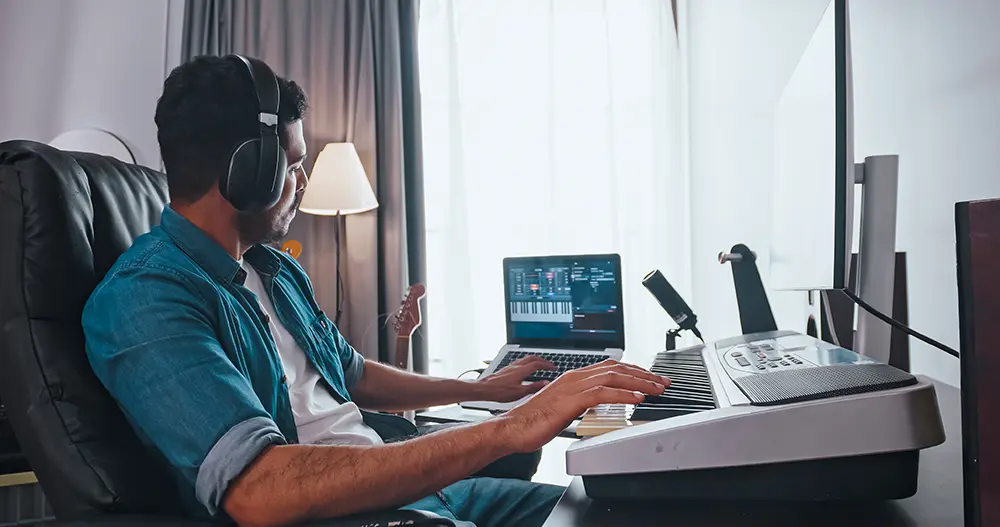 Man sitting at desk playing keyboard looking at laptop and music software