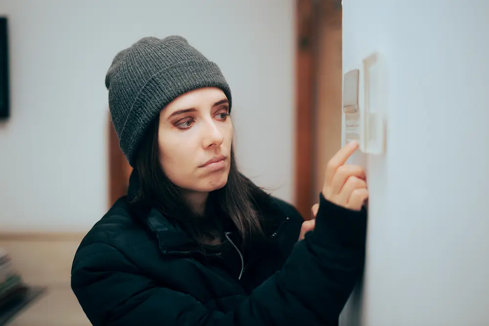 A woman in a sweater and winter hat adjusts the thermostat in cold house